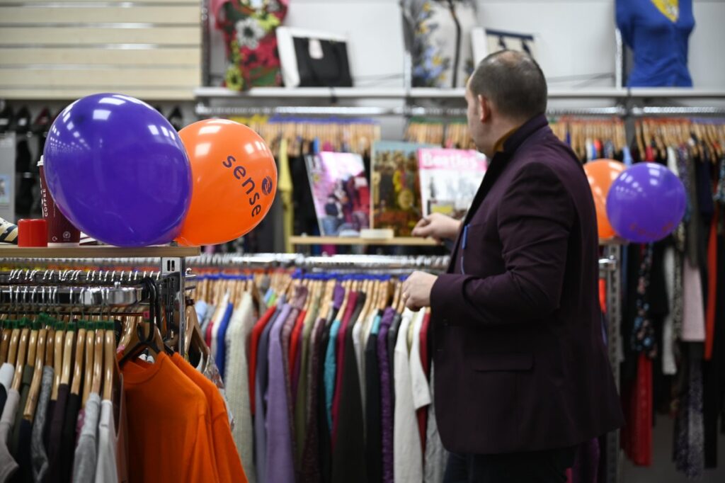 Clothes on racks and Sense balloons, as a man looks in the charity shop