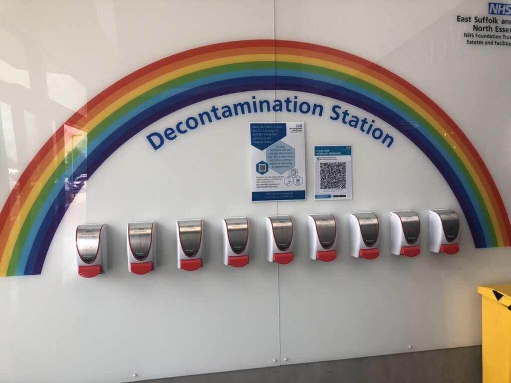 Row of nine hand sanitisers under a painted rainbow at the entrance to Colchester hospital. Photo: Howard Lake, 2021