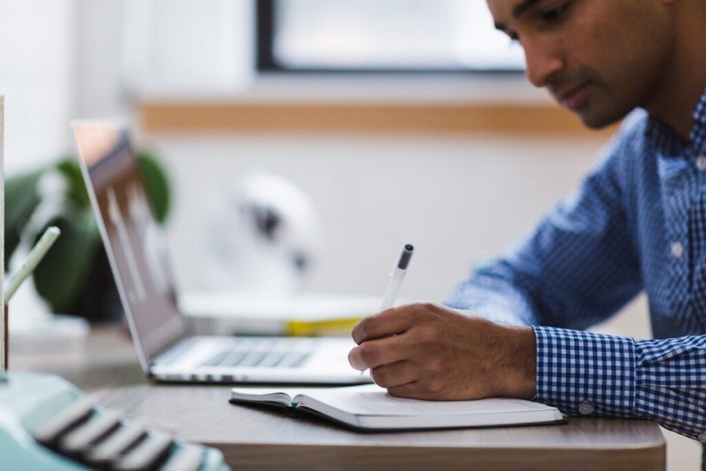 man writing on a notepad by a computer