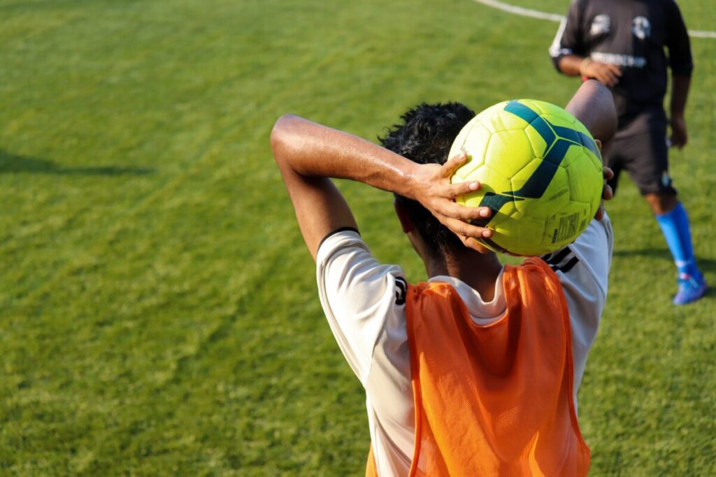 A young man in an orange vest holds a yellow football behind his head ready to throw