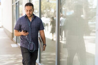 Man in tshirt walking down a sunny street, listening to a podcast on his phone. Image: Pexels.com