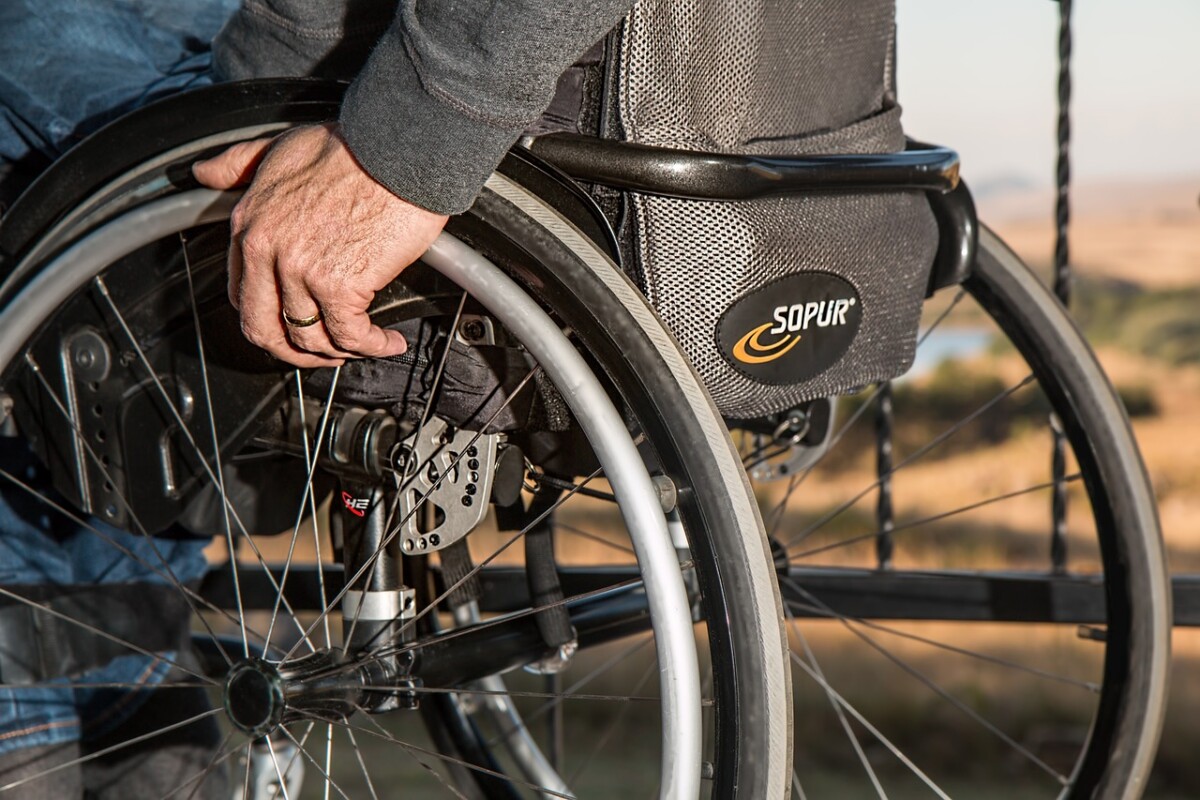 The wheels of a wheelchair with a man's hand propelling them forward. Image by Steve Buissinne from Pixabay
