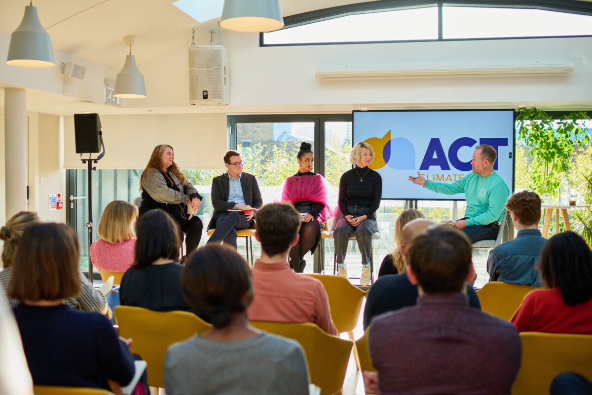 Photo of discussion panel seated in front of a screen with 'ACT' on it. Taken from the rear of a room with delegates sitting in rows. Left to right_Louise Brown (Supporter Relations Coordinator, Friends of the Earth), Christopher Hammond (Chief Executive, UK100), Hirra Khan Adeogun (Co-Director, Possible), Harriet Kingaby (Head of ACT Climate Labs), Jake Dubbins, Managing Director of Media Bounty