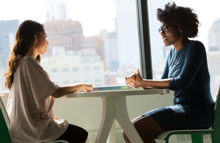 two women sit opposite each other at a small table in a window. By Christina Wocintechchat on unsplash