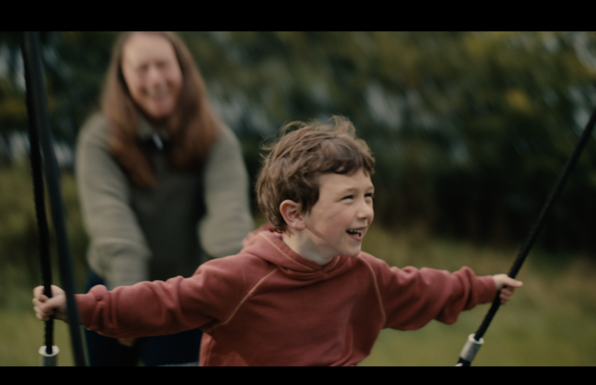 A smiling child on a swing, with his mother, out of focus, behind him, smiling too. Image: CHAS