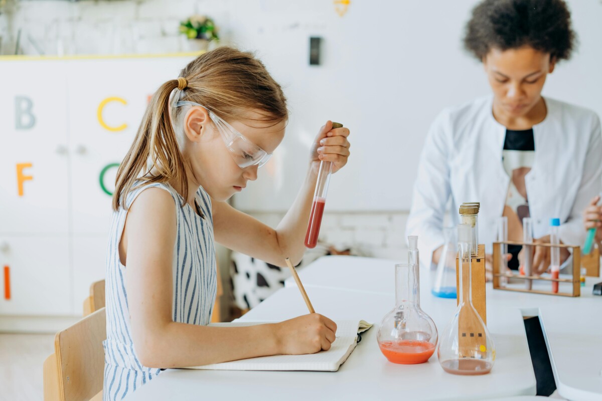 A young woman in lab coat and safety glasses looks at a small beaker of liquid. By Mikhail Nilov on Pexels