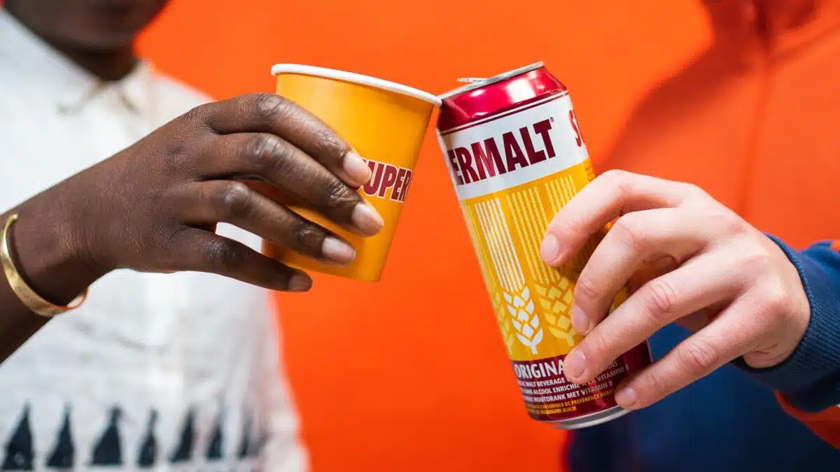 Supermalt can and branded paper cup being held by two people.