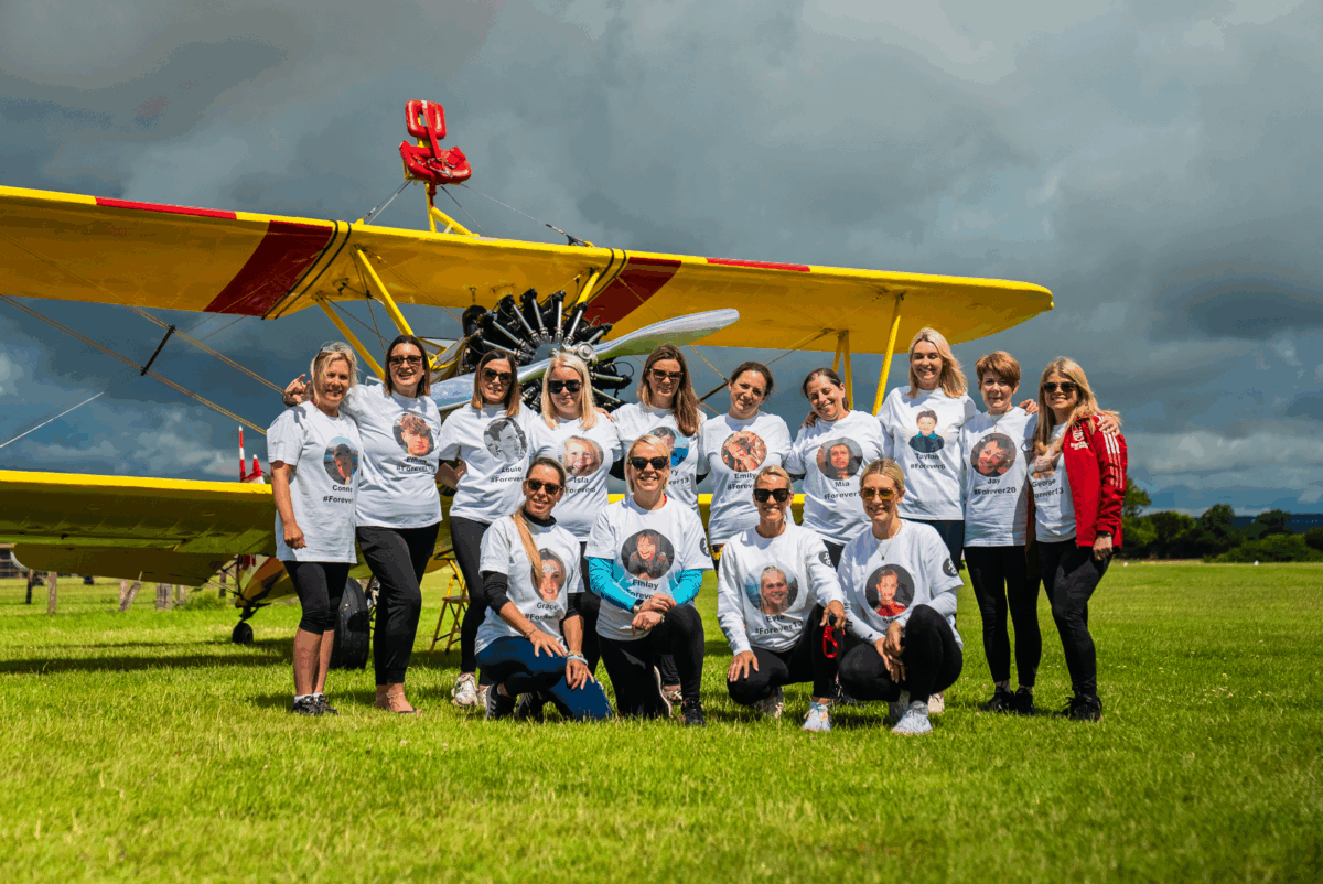 Angel Mums lined up in front of a wing-walking bi-plane on a grass runway.