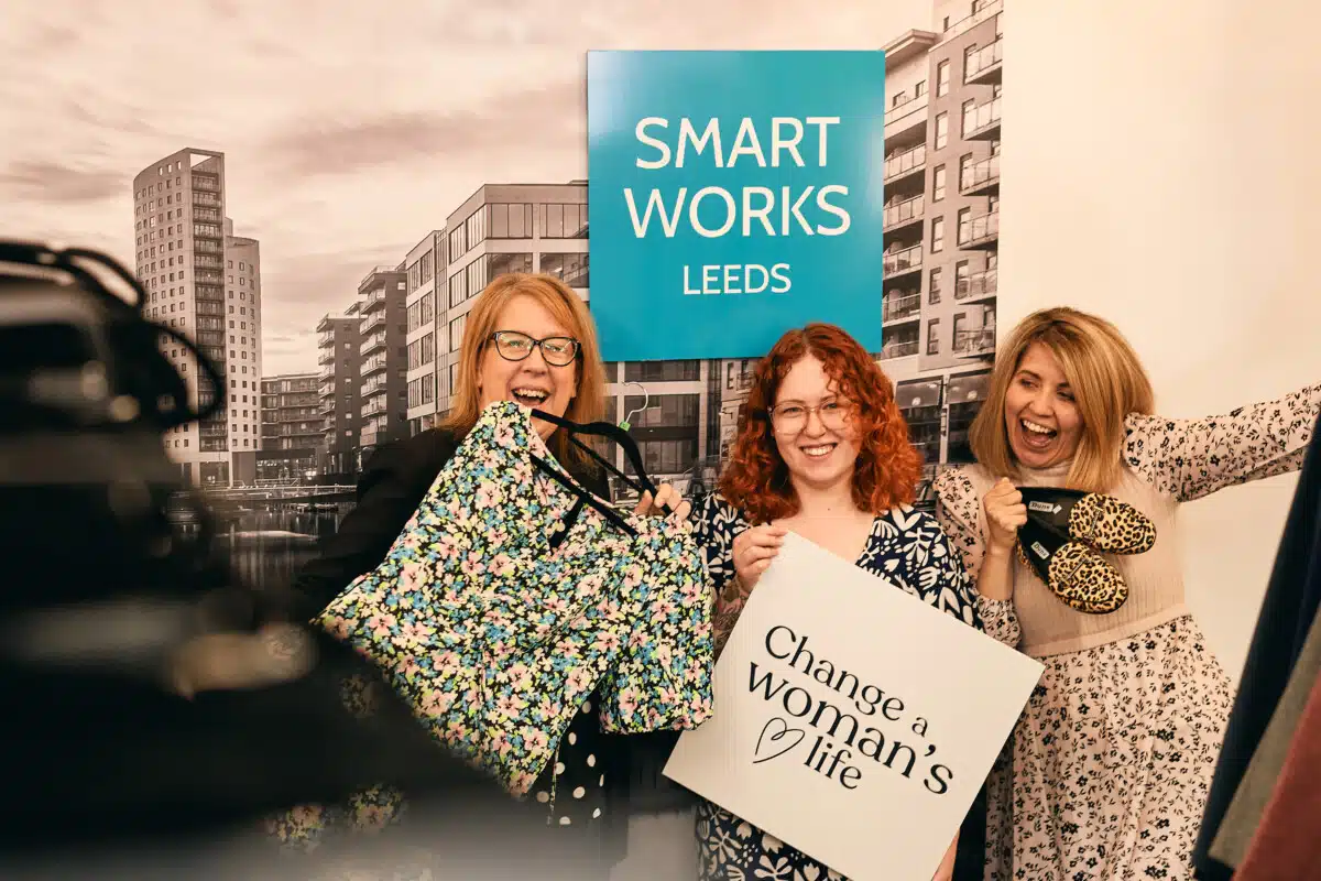 Three women celebrating holding shopping bags with 'Change a Woman's Life" on one of them, in front of a teal-coloured "Smart Works Leeds' sign.