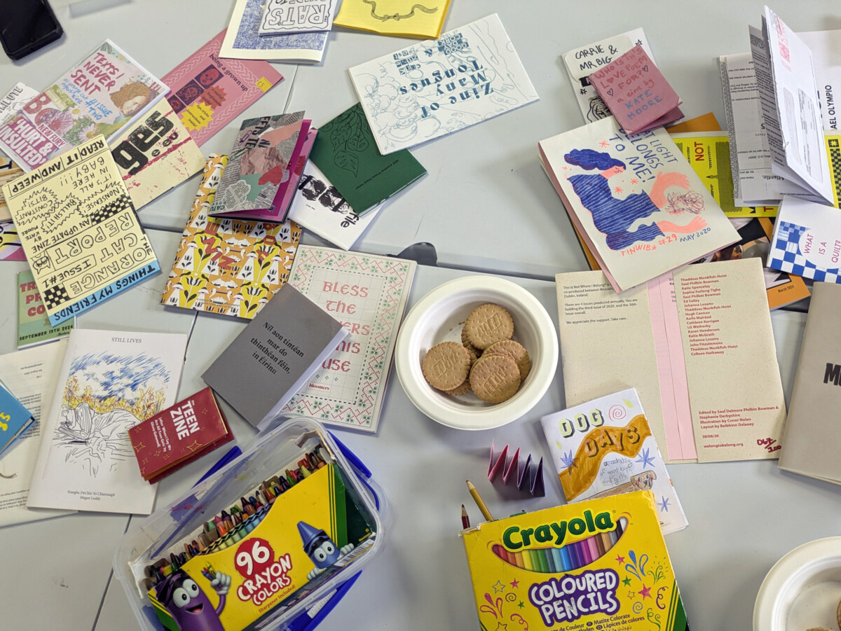 Zines workship at Graffiti Theatre, Cork. Zines, biscuits, crayons and other creative items are scattered on a table and viewed from above.
