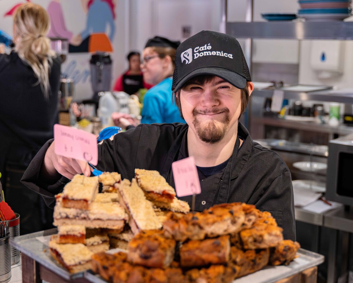 A smiling young person working in Cafe Domenica, standing behind a tray full of cakes.