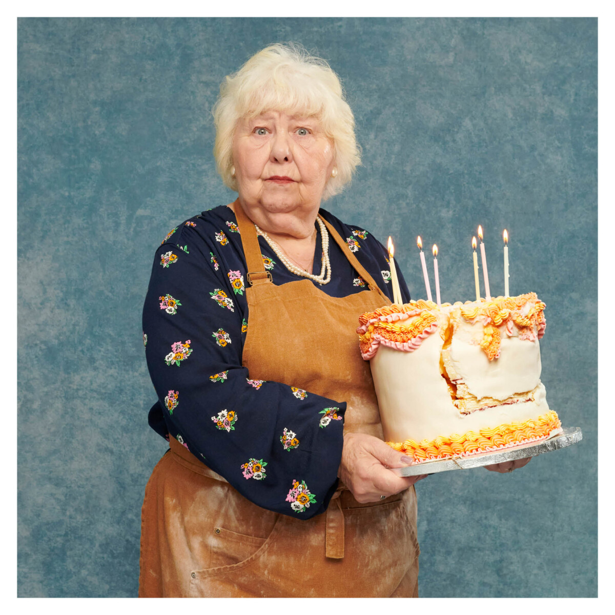 A grey-haired older woman in a pinafore holds a giant birthday cake with candles, against a plain background