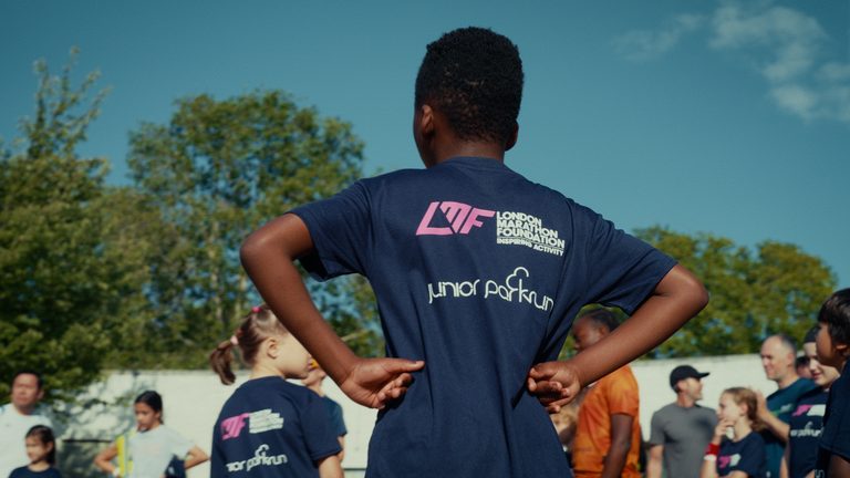 A child with arms on hips viewed from behind at a junior parkrun on a sunny morning. He wears a navy blue London Marathon Foundation tshirt.