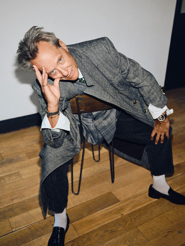 RIchard E Grant, sitting, head on one hand, in a grey coat, black trousers, and unbuttoned shirt sleeves.